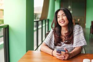 leende ung asiatisk kvinna bär klänning och känsla Lycklig, Sammanträde på en restaurang för en frukost medan innehav en smartphone foto