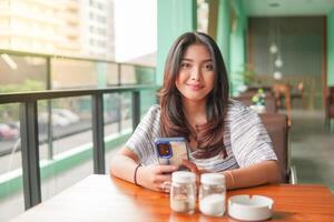 leende ung asiatisk kvinna bär klänning och känsla Lycklig, Sammanträde på en restaurang för en frukost medan innehav en smartphone foto