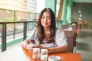 ung asiatisk kvinna bär klänning Sammanträde på en restaurang för en frukost, ser på smartphone i en djup trodde med allvarlig ansikte foto