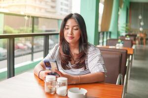 ung asiatisk kvinna bär klänning Sammanträde på en restaurang för en frukost, ser på smartphone i en djup trodde med allvarlig ansikte foto