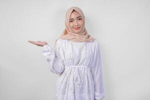 leende asiatisk kvinna bär hijab och vit klänning pekande upp till de kopia Plats på henne rätt sida, stående över isolerat vit bakgrund. ramadan och eid mubarak annons begrepp foto