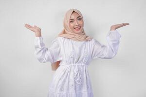Lycklig asiatisk kvinna bär hijab och vit klänning pekande upp och presenter de alternativ på kopia Plats ovan, stående över isolerat vit bakgrund. ramadan och eid mubarak annons begrepp foto