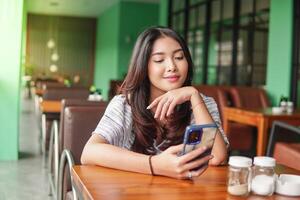 dagdrömma ung asiatisk kvinna bär klänning Sammanträde på en restaurang för en frukost, innehav en smartphone medan stöttning henne haka förlorat i trodde foto