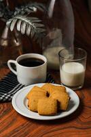 knaprig småkakor kex eras i tallrik med svart kaffe och glas av mjölk isolerat på tabell sida se av amerikan Kafé bakad mat foto