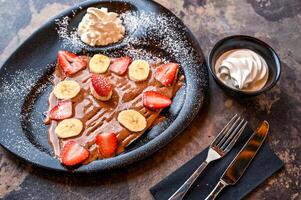 jordgubb och banan crepe med choklad, vispad grädde, kniv och gaffel eras i maträtt isolerat på mörk bakgrund närbild topp se av Kafé bakad efterrätt mat foto