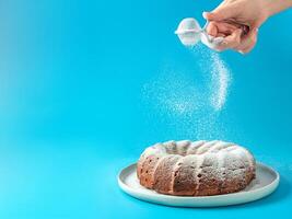 kvinnas hand stänk glasyr socker över färsk Hem tillverkad bundt kaka. pulver socker falls på färsk perfekt bunt kaka över blå bakgrund. kopia Plats för text. idéer och recept för frukost eller efterrätt foto