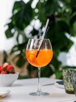 sommar dryck aperol spritz aperitif eras i vin glas med aperol, prosecco, soda, skiva av orange. uppfriskande dryck aperol spritz cocktail på tabell i interiör med grön monstera växt bakgrund foto