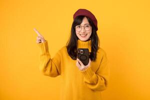 porträtt ung skön 30s asiatisk kvinna Lycklig leende bär gul skjorta och röd hatt pekande finger gest till fri kopia Plats och använder sig av smartphone mot gul bakgrund. ny Ansökan foto