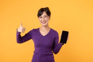 upphetsad ung asiatisk kvinna 30s bär lila skjorta och glasögon som visar mobil telefon skärm, tummen upp, tycka om smth Bra, rekommenderar smartphone Ansökan, stående över gul bakgrund foto