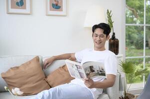 Lycklig asiatisk man Sammanträde på soffa läsning en bok i levande rum på Hem, koppla av tid och livsstil begrepp foto