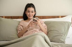 ung asiatisk kvinna använder sig av smart telefon liggande på säng foto