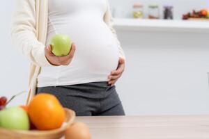 gravid kvinna innehav ett äpple i främre av en tabell med en skål av frukt. scen är positiv och friska, som de kvinna är välja till äta en näringsrik, hälsa vård foto