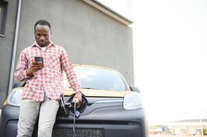 tillfällig afrikansk man med smartphone nära elektrisk bil väntar för de Avsluta av de batteri laddning bearbeta. foto