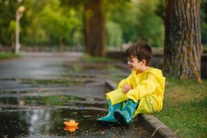 barn spelar med leksak båt i pöl. unge spela utomhus- förbi regn. falla regnig väder utomhus aktivitet för ung barn. unge Hoppar i grumlig pölar. vattentät jacka och stövlar för bebis. barndom foto