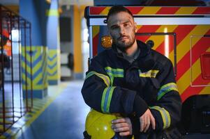 porträtt av en brandman i en skyddande kostym och en skyddande hjälm stående förbi en brand motor efter arbetssätt på en brand. närbild bild foto