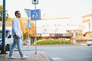 ung blind man med vit sockerrör gående tvärs över de gata i stad foto