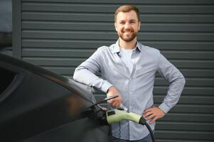 stilig skäggig man i tillfällig ha på sig, stående på de laddning station och laddare för ett elektrisk bil. eco elektrisk bil begrepp foto