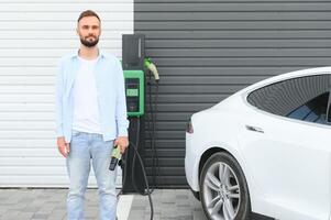 skön ung eleganta man är med elektrisk bil på dagtid laddning de fordon foto