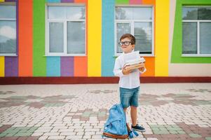 tillbaka till skola. Lycklig leende pojke i glasögon är gående till skola för de först tid. barn med ryggsäck och bok utomhus. början av lektioner. först dag av falla. foto