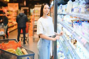 ung leende Lycklig kvinna 20s ha på sig tillfällig kläder handla på supermarket Lagra köpa välja mjölk foto