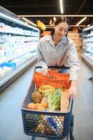 ung nöjd roligt kvinna i tillfällig kläder handla på supermarket Lagra med matvaror vagn foto