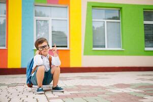 ledsen liten pojke utanför av skola. ledsen skol med böcker nära en modern skola. skola begrepp. tillbaka till skola. foto