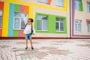 tillbaka till skola. Lycklig leende pojke i glasögon är gående till skola för de först tid. barn med ryggsäck och bok utomhus. början av lektioner. först dag av falla foto