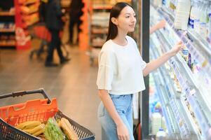 ung leende Lycklig kvinna 20s i tillfällig kläder handla på supermarket Lagra med matvaror vagn foto