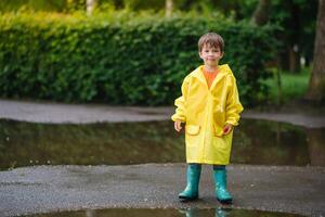 liten pojke spelar i regnig sommar parkera. barn med paraply, vattentät täcka och stövlar Hoppar i pöl och lera i de regn. unge gående i sommar regn utomhus- roligt förbi några väder. Lycklig barndom foto