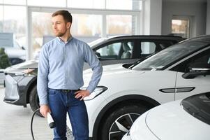 skägg man påfrestande en ny laddning kabel- med en bil laddning station på de motor återförsäljare. begrepp av uppköp elektrisk fordon. smart ekologisk levande foto