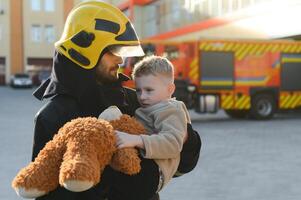en brandman ta en liten barn pojke till spara honom. brand motor bil på bakgrund foto