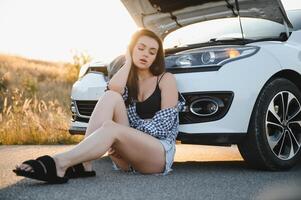 en ung flicka sitter nära en bruten bil på de väg med ett öppen huva. foto