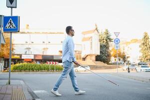 ung blind man med vit sockerrör gående tvärs över de gata i stad foto