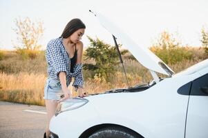 attraktiv smal ung flicka i sommar shorts och skjorta reparationer en bruten bil. en skön kvinna står nära Uppfostrad bil huva. foto