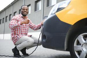 afrikansk man innehav avgift kabel- i på hand stående nära elektrisk bil foto