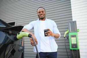 eleganta afrikansk man med coffe kopp i hand skär plugg in i de elektrisk bil laddning uttag foto