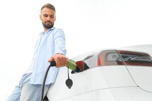 ung stilig man innehav laddning kabel- på elektrisk laddning station punkt stående nära hans ny bil foto