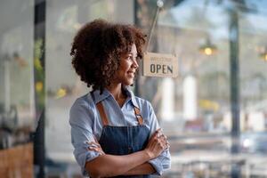 kvinna afrikansk kaffe affär små företag ägare bär förkläde stående och se utanför de affär, i de bakgrund där är en Välkommen tecken foto