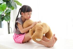 söt liten asiatisk flicka har roligt hon kramade en stor brun teddy Björn på de vit säng. begrepp av familj, vänskap foto