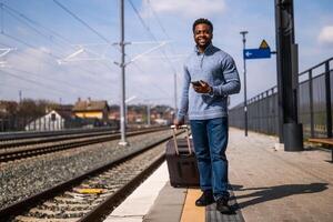 Lycklig man med en resväska använder sig av telefon medan gående på de järnväg station. foto