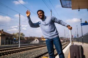 Lycklig man med resväska stående och vinka på en järnväg station. foto