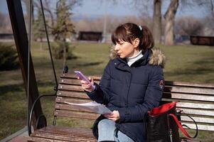 en ung kvinna revisor sitter på en parkera bänk och läser en rapportering dokumentera och samtal på en smartphone, rapportering period foto
