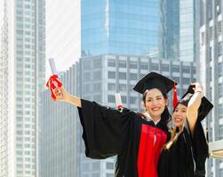 två lycka graderad kvinna i svart gradering kappor innehav diplom och kramas varje Övrig på gradering ceremoni, stående utomhus med stad byggnader bakgrund foto