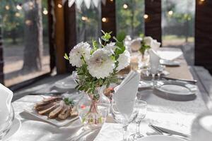 en lång festlig tabell dekorerad med vit blommor och grönska. där är tallrikar, glasögon och ljus på de tabell. de rum är dekorerad med vit girlanger. foto