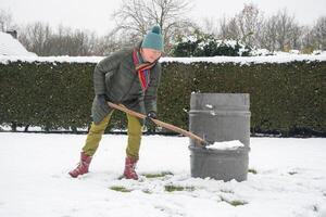 mitten åldrig kvinna är samlar snö i en tunna med en spade, naturlig Resurser foto