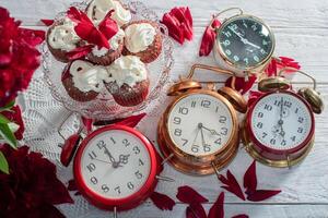 samling av årgång klockor på de tabell, röd sammet muffin på en tallrik, scharlakansrött skum kronblad på en vit bordsduk foto