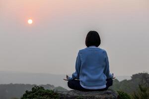 tillbaka se av kvinna är avkopplande praktiserande meditation yoga mudra på berg topp med stigande Sol under sommar till uppnå lycka från inre fred visdom för friska sinne och själ foto