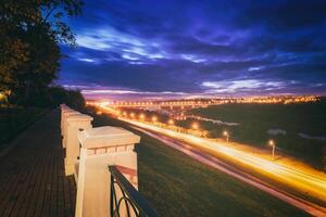 rör på sig bil med fläck ljus genom stad på natt. bro över de flod och de väg. en se från de parkera från en höjd med en staket i de förgrund. årgång filma estetisk. foto