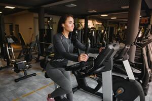 ung kvinna med hörlurar håller på med övningar på brevpapper cykel i en Gym eller kondition Centrum. ung sportig kvinna i Gym lyssna musik från smartphone. kvinnor håller på med konditionsträning övningar foto