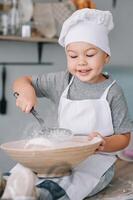 ung pojke söt på de kök laga mat kock i vit enhetlig och hatt nära tabell. hemlagad pepparkaka. de pojke kokta de choklad småkakor foto